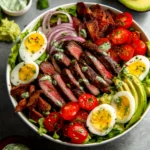 Close-up of a loaded steak Cobb salad with sliced steak, boiled eggs, avocado, cherry tomatoes, bacon, and creamy dressing in a bowl